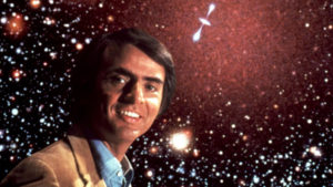 The late Carl Sagan looks at the viewer. The background image is of outer space, densely populated with stellar objects.
