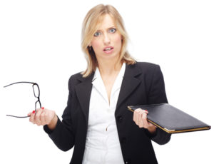 A woman dressed in office attire holds her glasses and a binder with a quizzical look on her face.