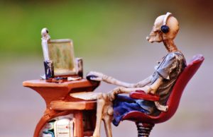 A sculpture of a skeleton wearing clothes and a headset. The skeleton is sitting at a desk using a computer.